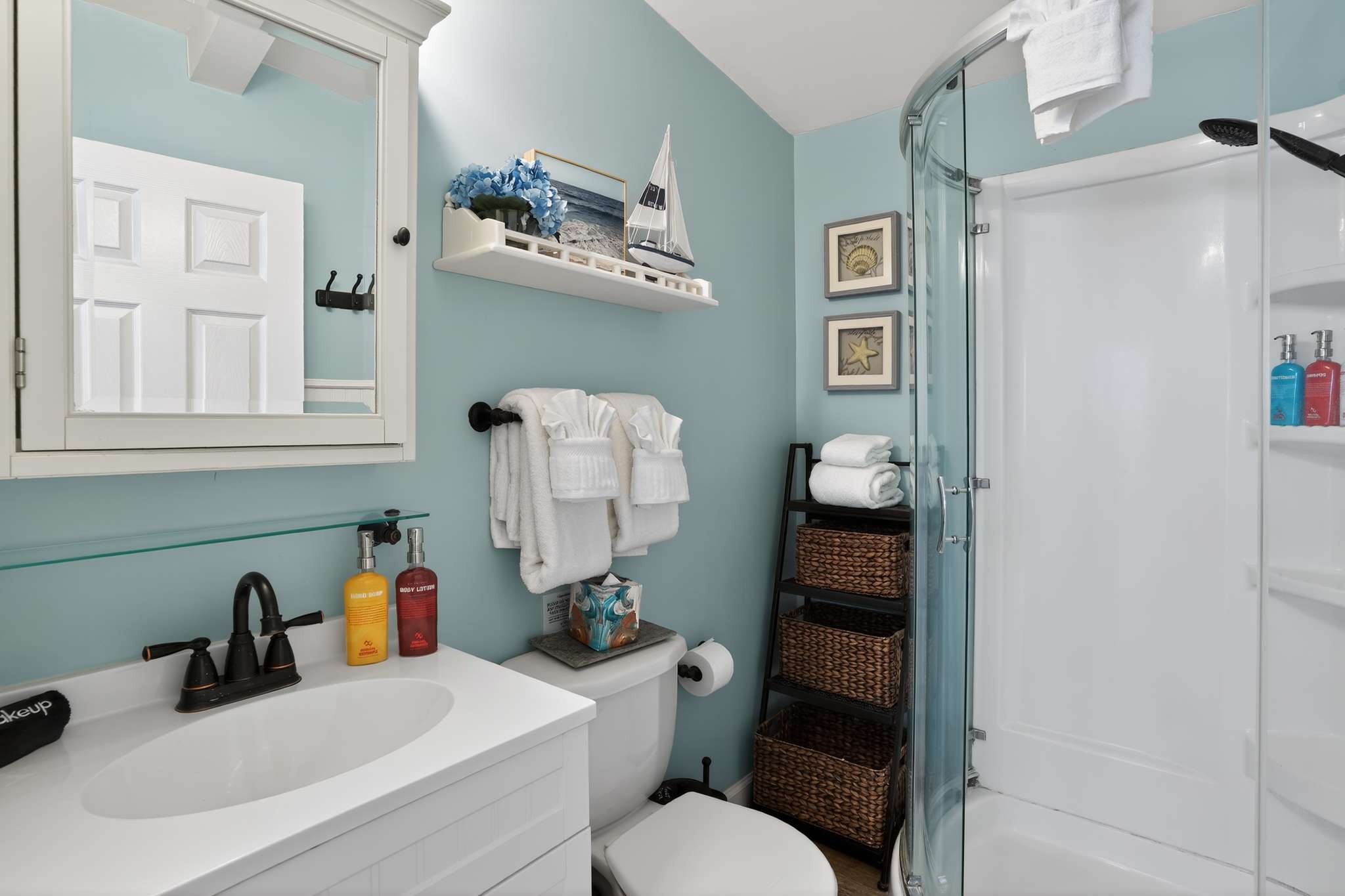 A compact bathroom with light blue walls and white wainscoting features a white vanity, a toilet, a corner glass shower, and decorative shelves with nautical accents.
