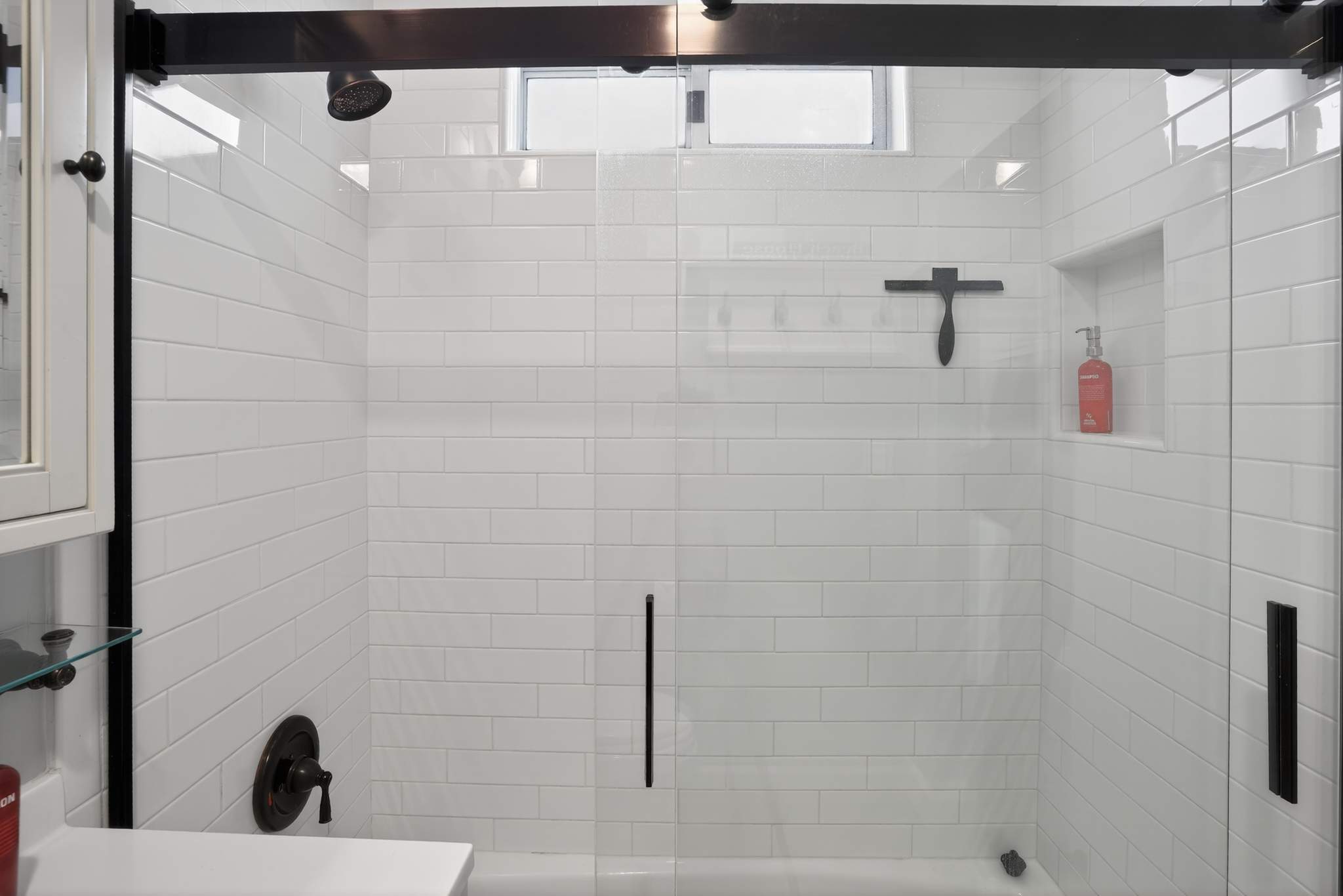 A close-up of a modern shower with white subway tiles, a dark bronze shower head, and a clear glass enclosure with black framing, featuring a built-in niche for toiletries.