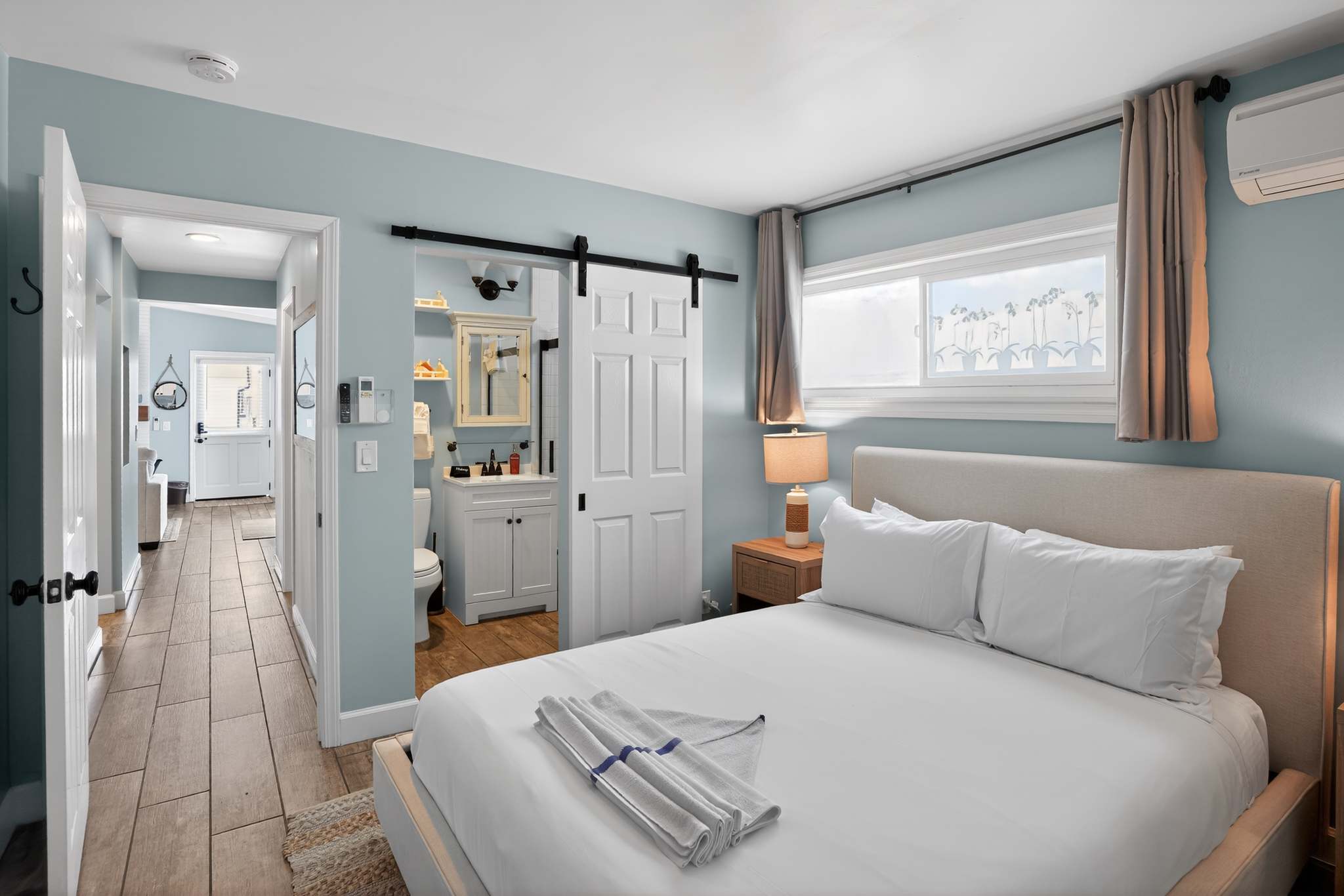 Serene bedroom with a light-colored upholstered king bed, light blue walls, and a white barn door that slides open to reveal a glimpse of a bathroom vanity.
