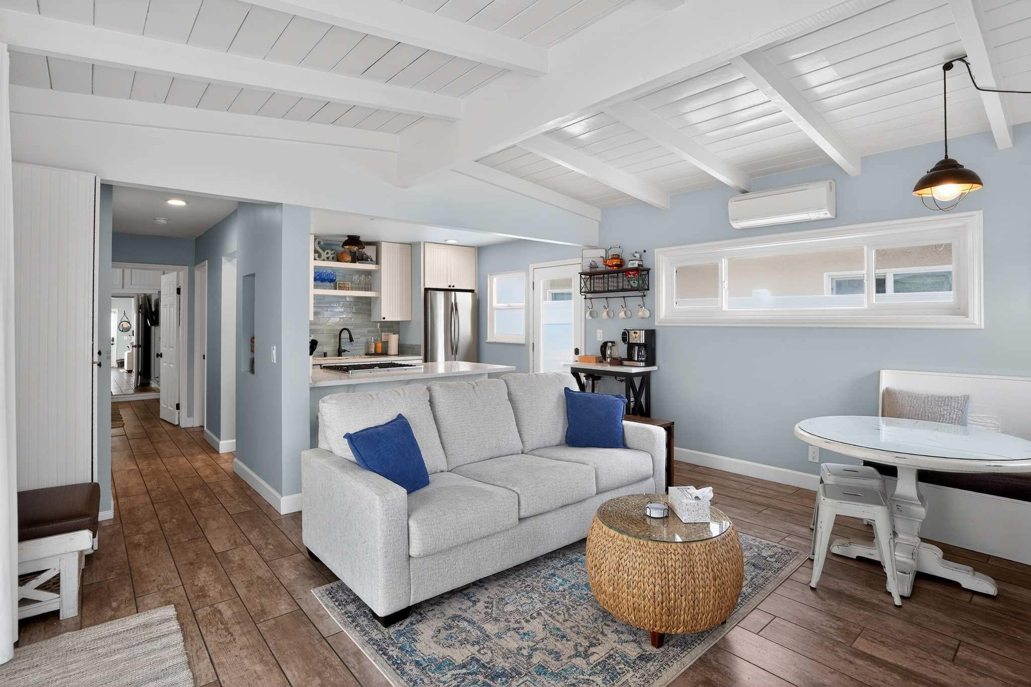 This open-concept living space features a light gray sofa with blue throw pillows, a round wicker coffee table, and wood-look tile flooring, leading into a bright kitchen and dining nook with light blue walls and a white beamed ceiling.