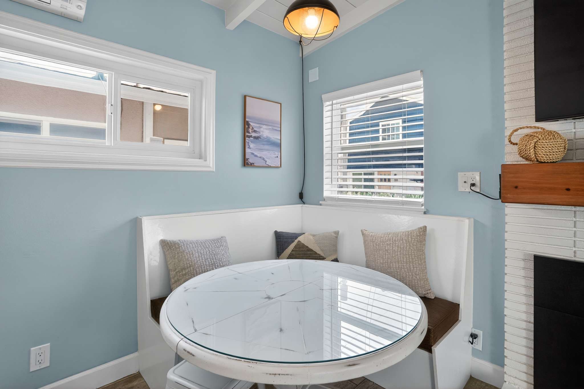 A charming built-in dining nook features a white banquette with brown cushions and throw pillows, surrounding a round white table with a glass top, set against light blue walls and wood-look tile flooring.