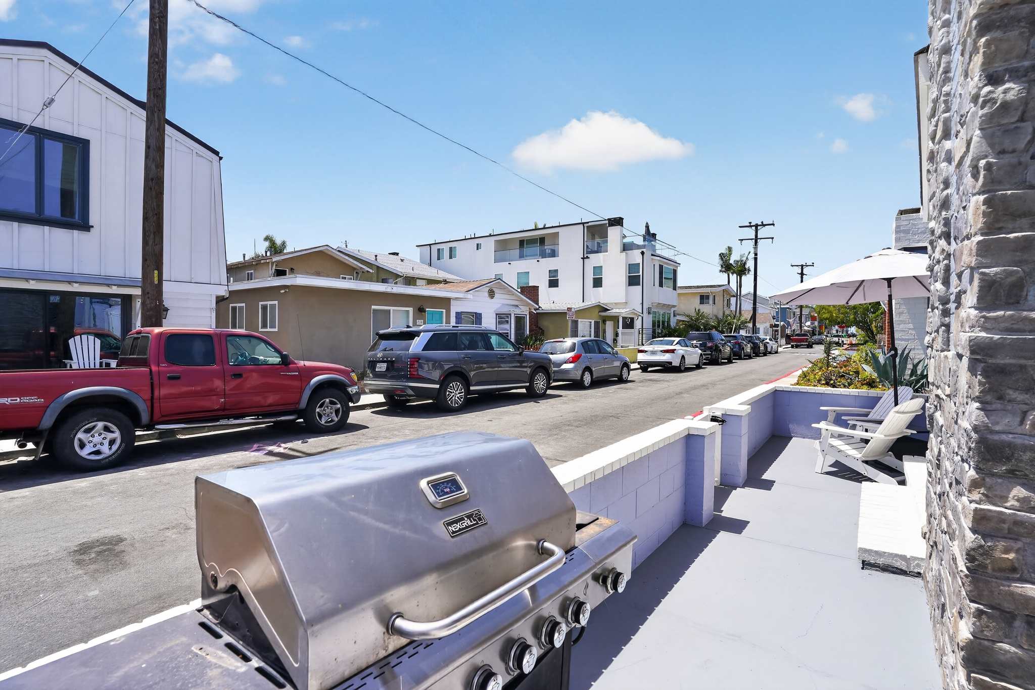 Street-level view of the property's front patio area, featuring a large stainless steel grill and a glimpse of the residential street with parked cars under a clear blue sky.