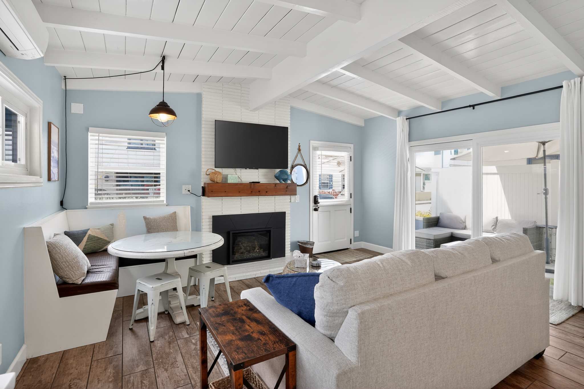 A cozy living room features a light gray sofa, a white brick fireplace with a mounted TV, and a built-in dining nook with a round white table, all under a white beamed ceiling and light blue walls.