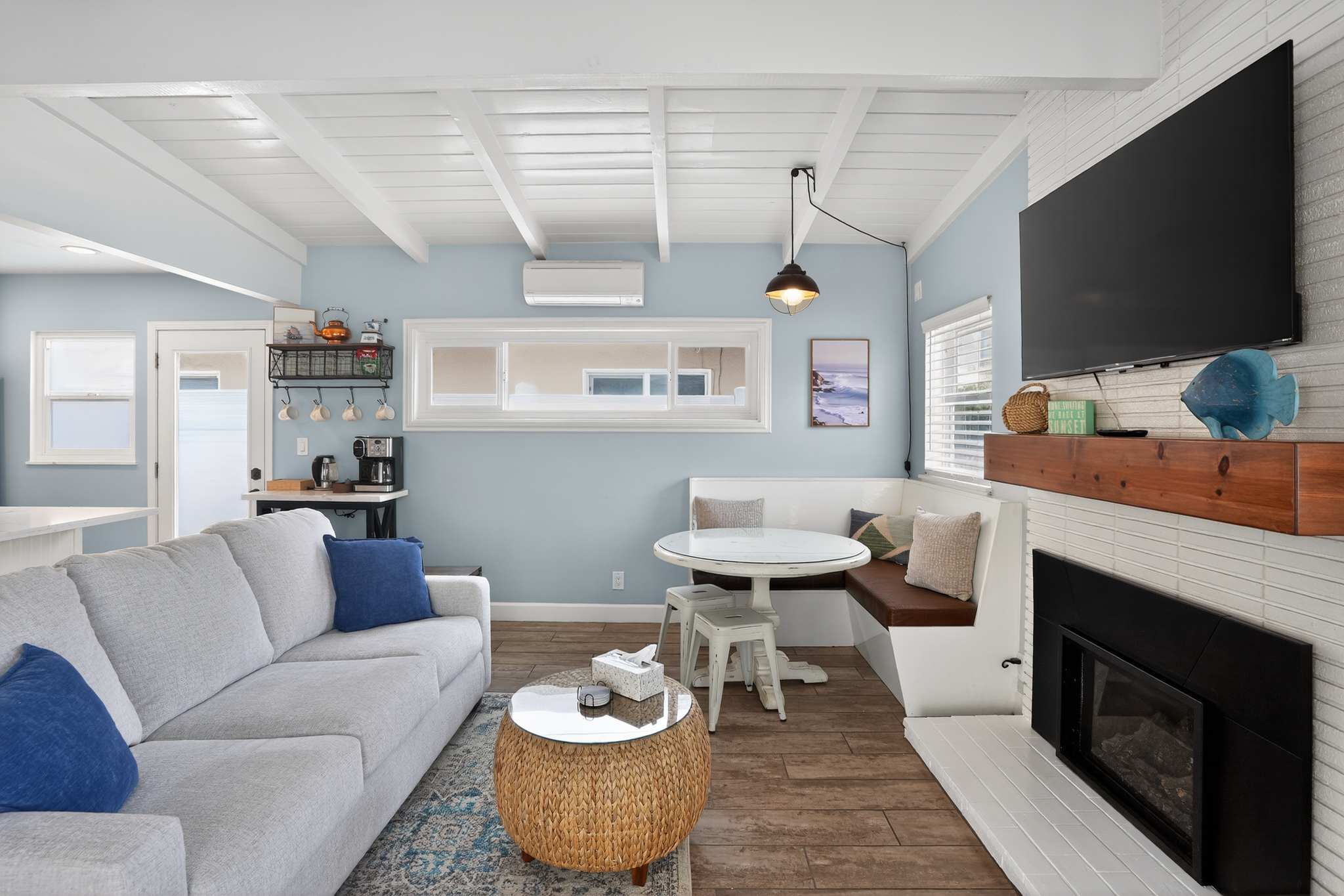 Cozy living room with light blue walls, exposed white beam ceiling, and wood-look tile flooring. A light gray sofa faces a fireplace with a mounted TV, and a built-in dining nook is to the right.