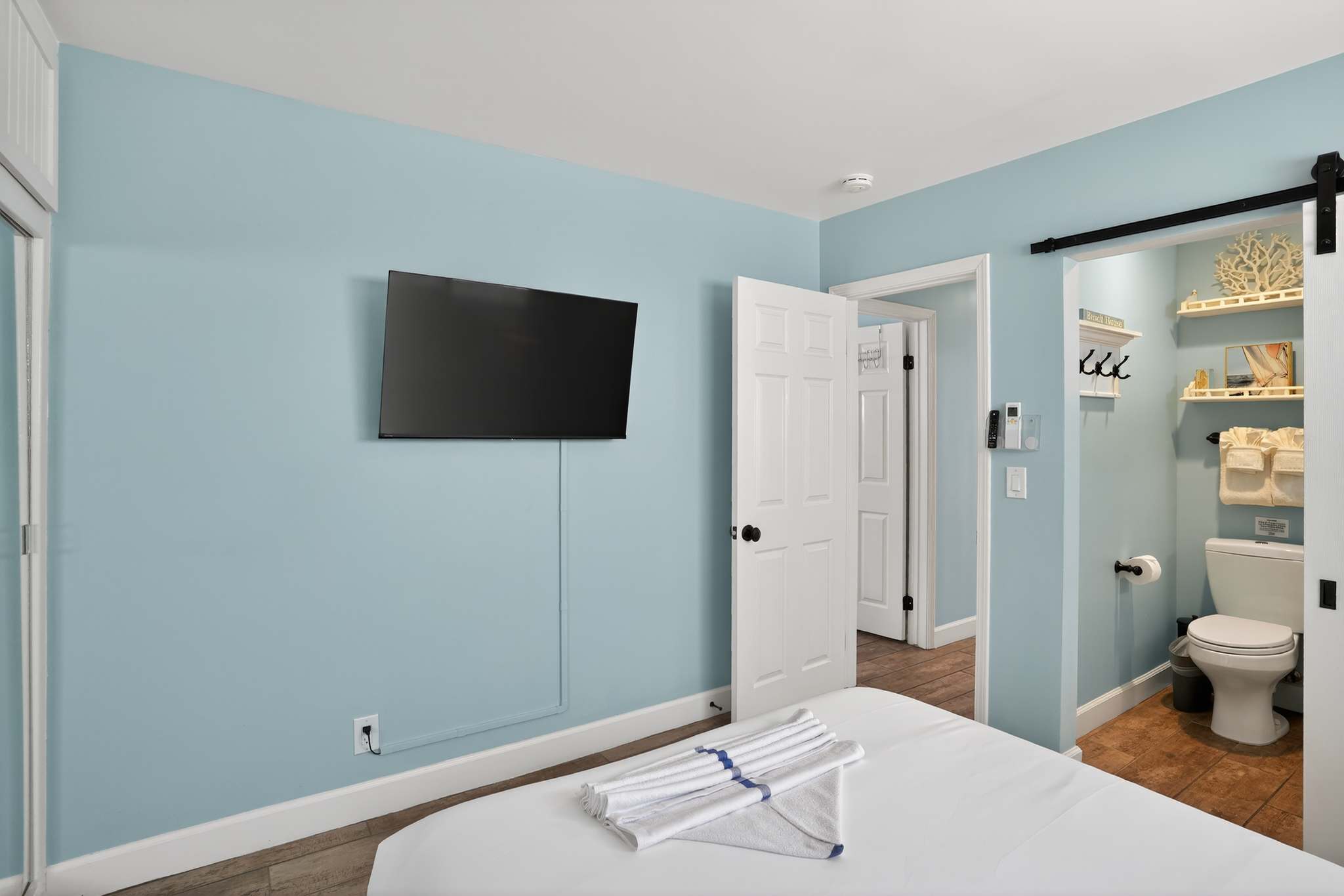 A bright bedroom with light blue walls features a white bed with folded towels, a wall-mounted flat-screen TV, and a partial view of a bathroom through a sliding barn door.