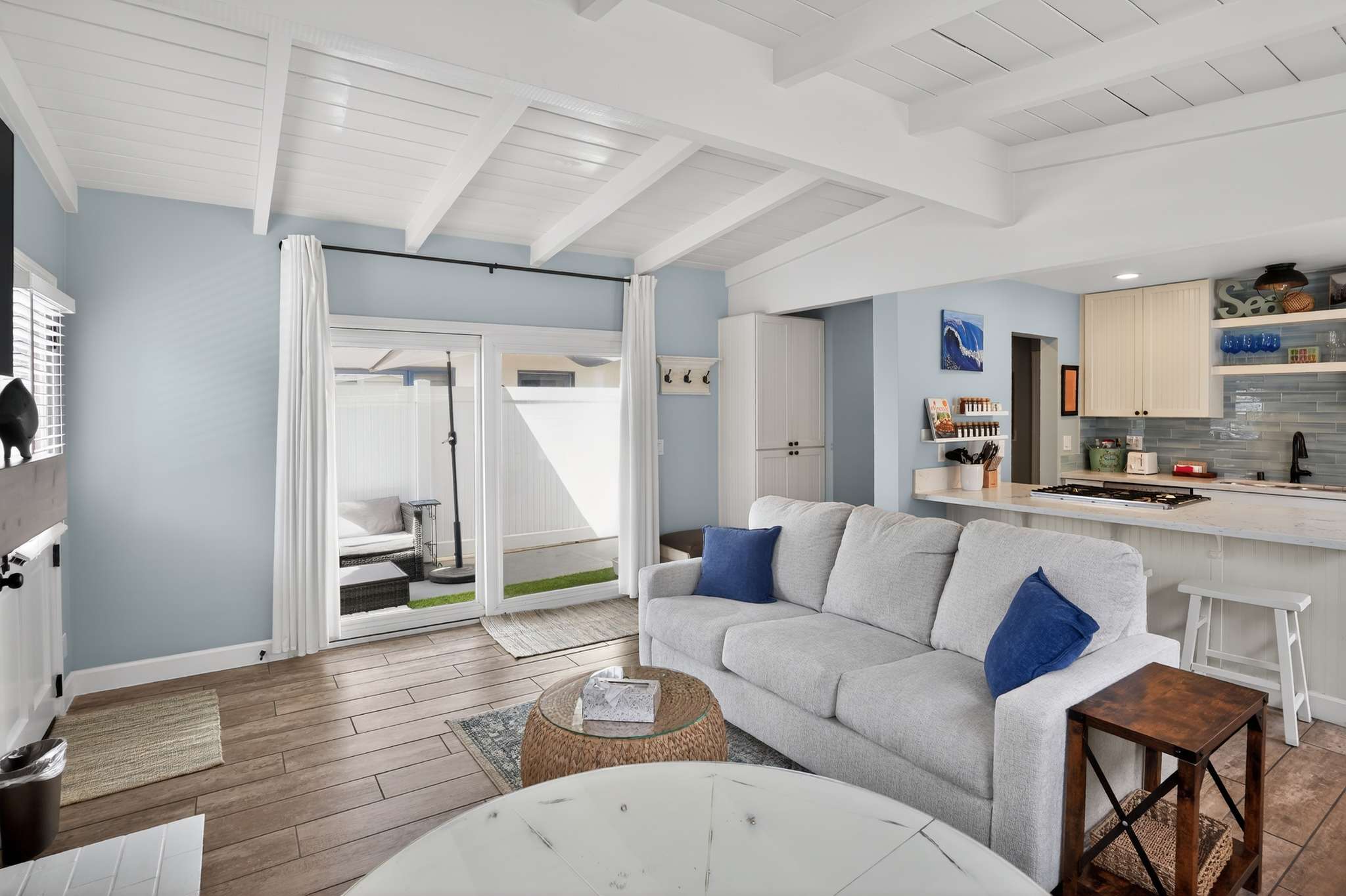 Bright living room with a light gray sofa, wood-look tile flooring, and an exposed white beam ceiling. A sliding glass door leads to an outdoor patio, and a coffee bar area is visible in the background.