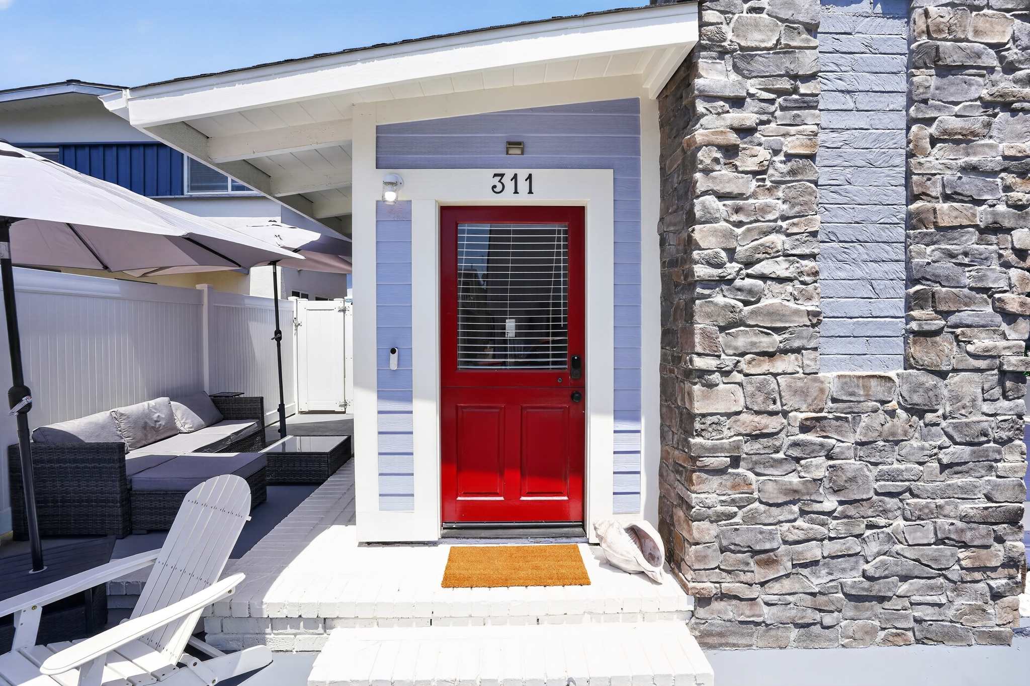 Front entrance of a light blue house with a vibrant red door, white trim, and stone siding. A small patio area with outdoor seating and umbrellas is visible to the left.