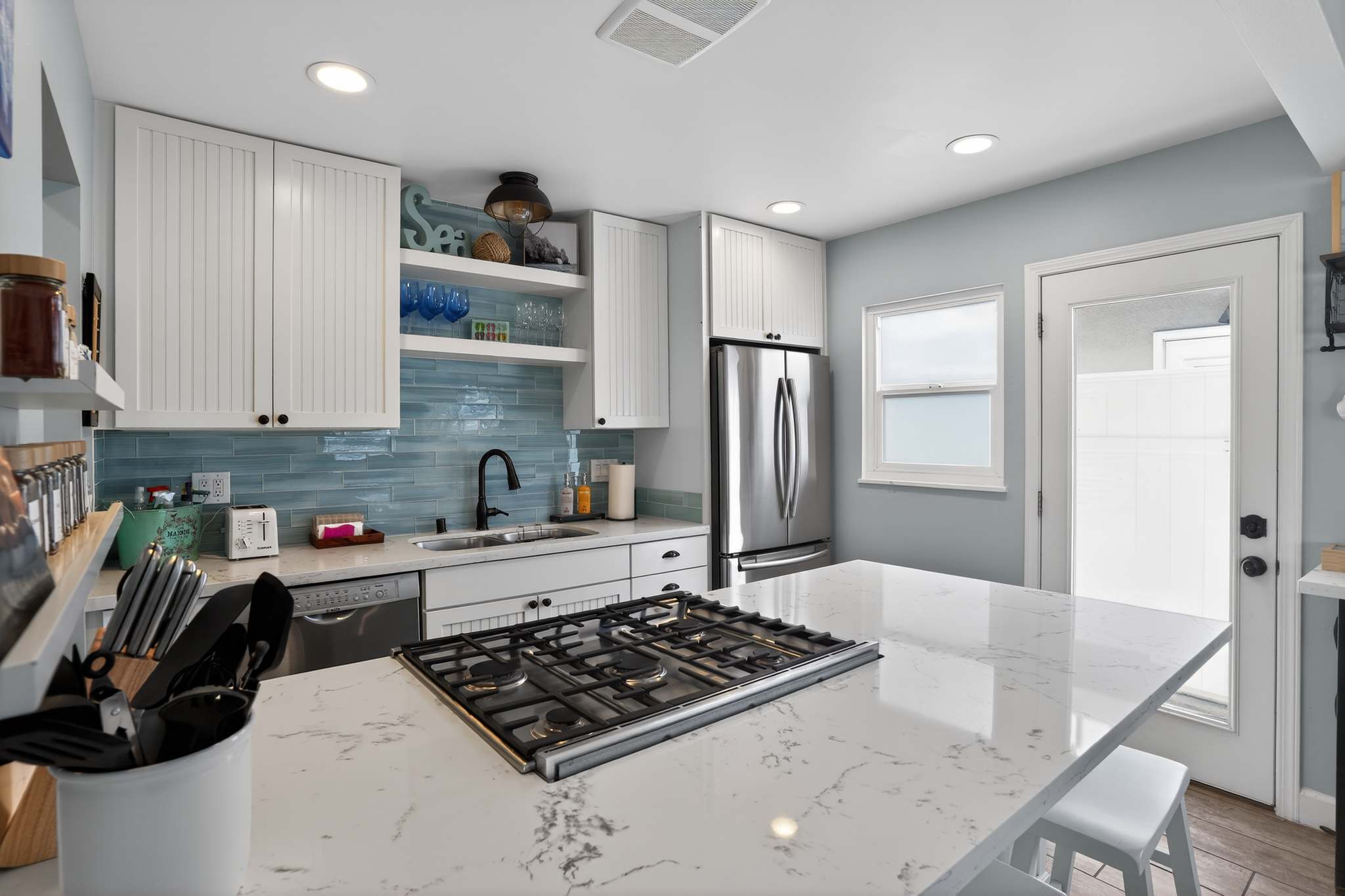 A bright kitchen with white shaker cabinets, light blue subway tile backsplash, and white quartz countertops, including a large island with a gas cooktop and bar seating, and a stainless steel French door refrigerator.