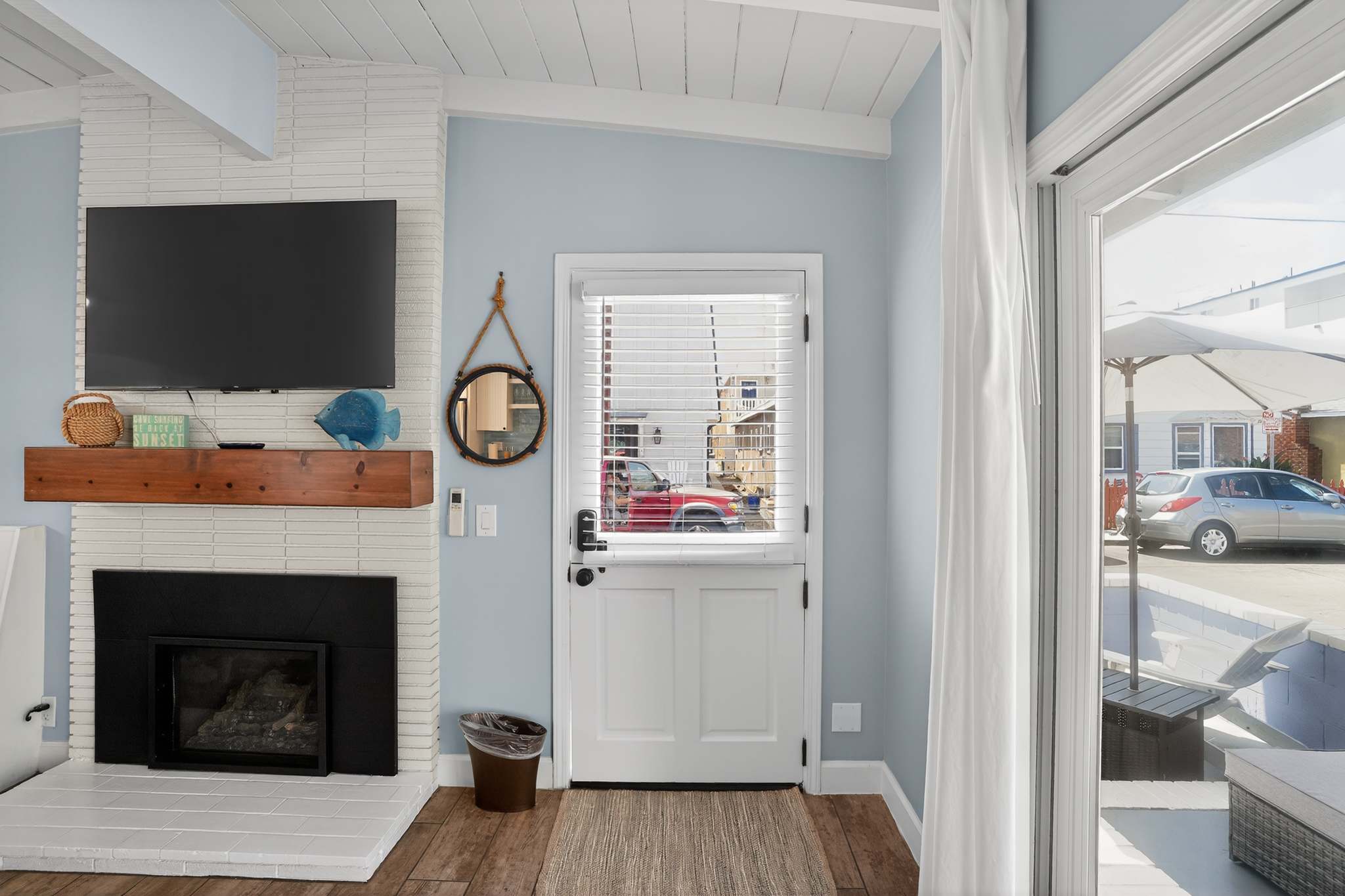 Detail shot of the living room's white brick fireplace with a rustic wooden mantel and a mounted TV, adjacent to a white Dutch door entrance with internal blinds.