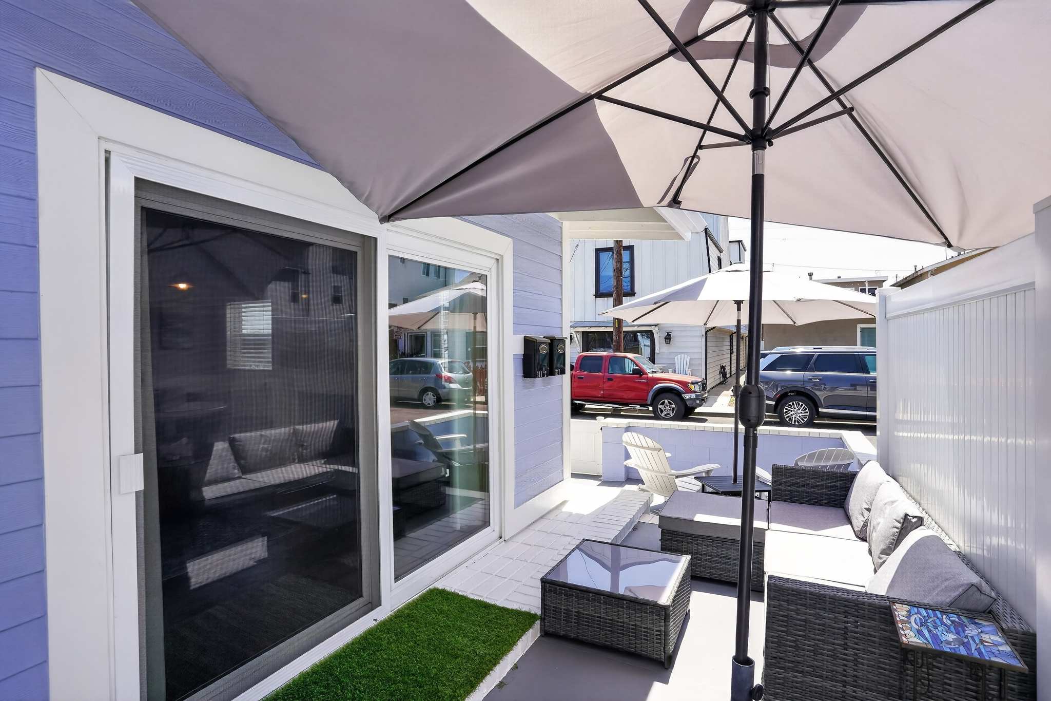 An inviting outdoor patio features comfortable wicker seating with grey cushions, a matching coffee table, and a large grey umbrella providing shade, with sliding glass doors leading indoors.