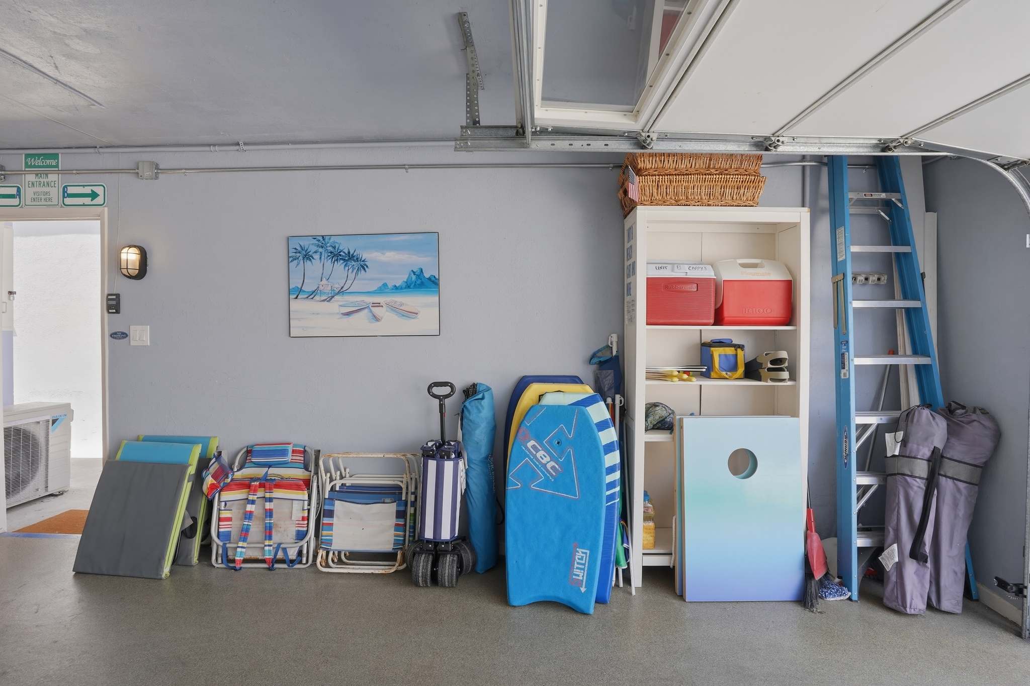 Garage interior showing various beach and recreational equipment, including boogie boards, beach chairs, and coolers, neatly stored against a light blue wall. A white shelving unit holds additional items.