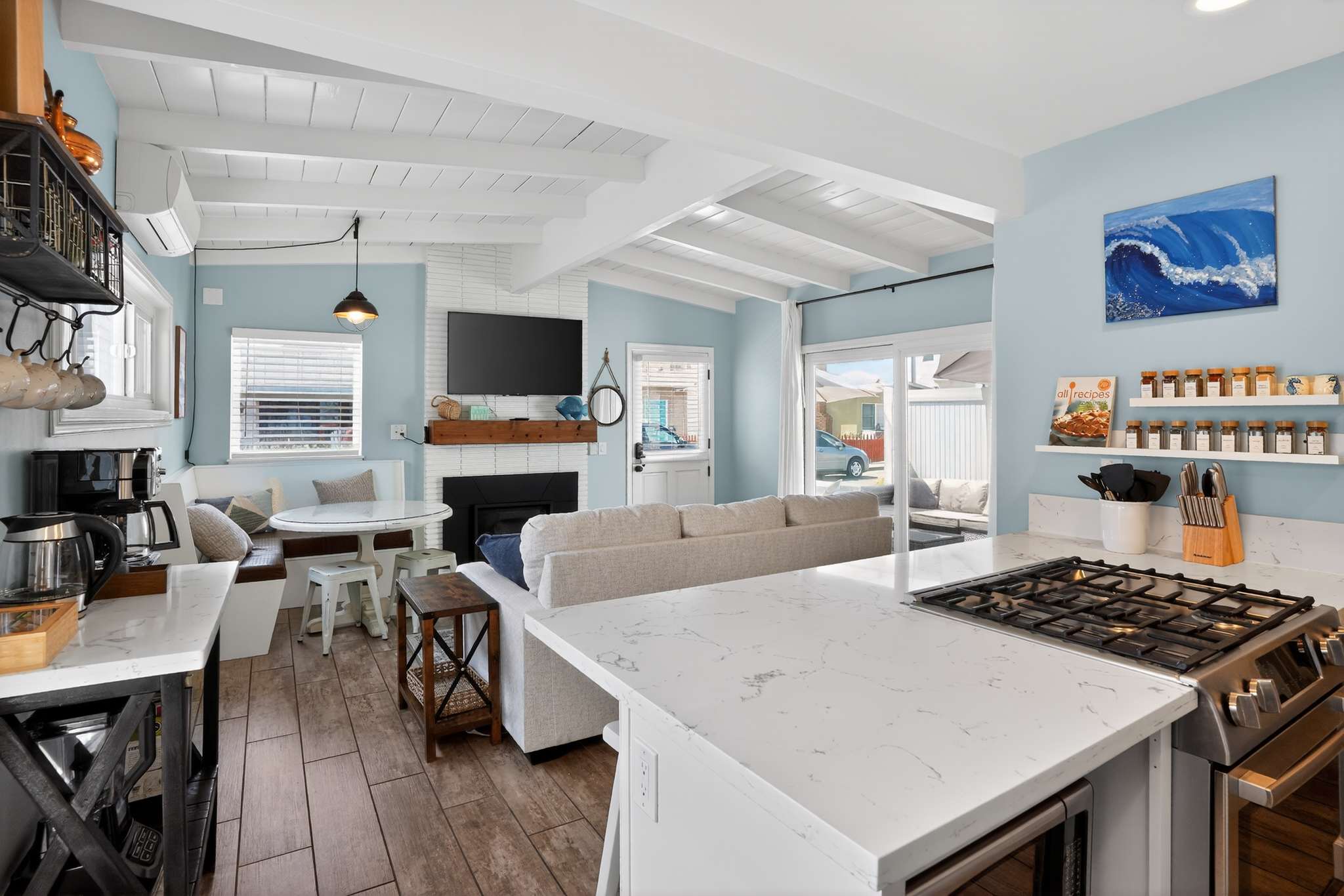 An expansive view from the kitchen island shows the open-concept living area with a light gray sofa, a white brick fireplace with a mounted TV, and a built-in dining nook, all under a white beamed ceiling.