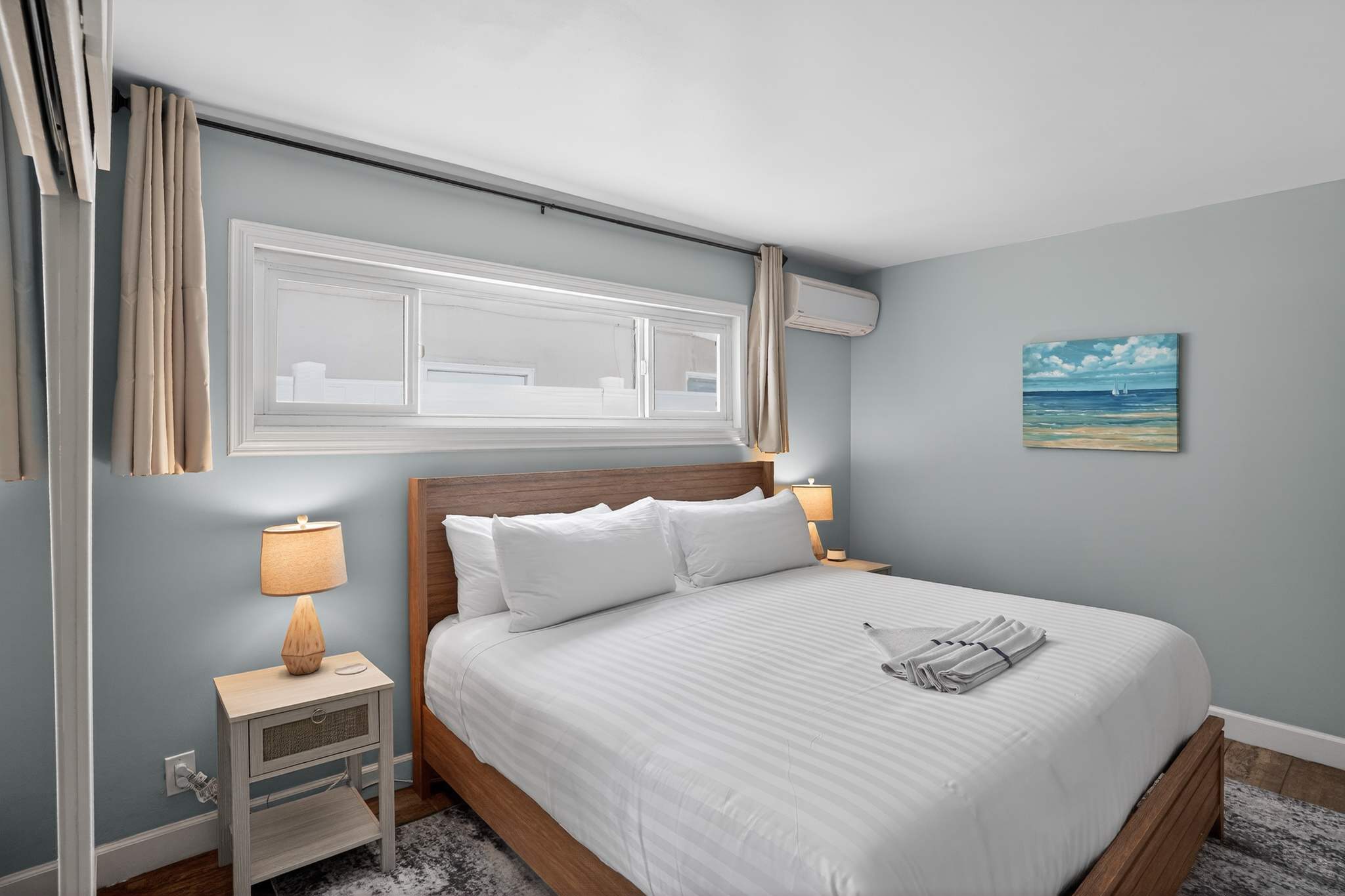 A bright bedroom with light blue walls features a wooden queen-sized bed with a light upholstered headboard, two light-colored nightstands with lamps, and a window above the bed.