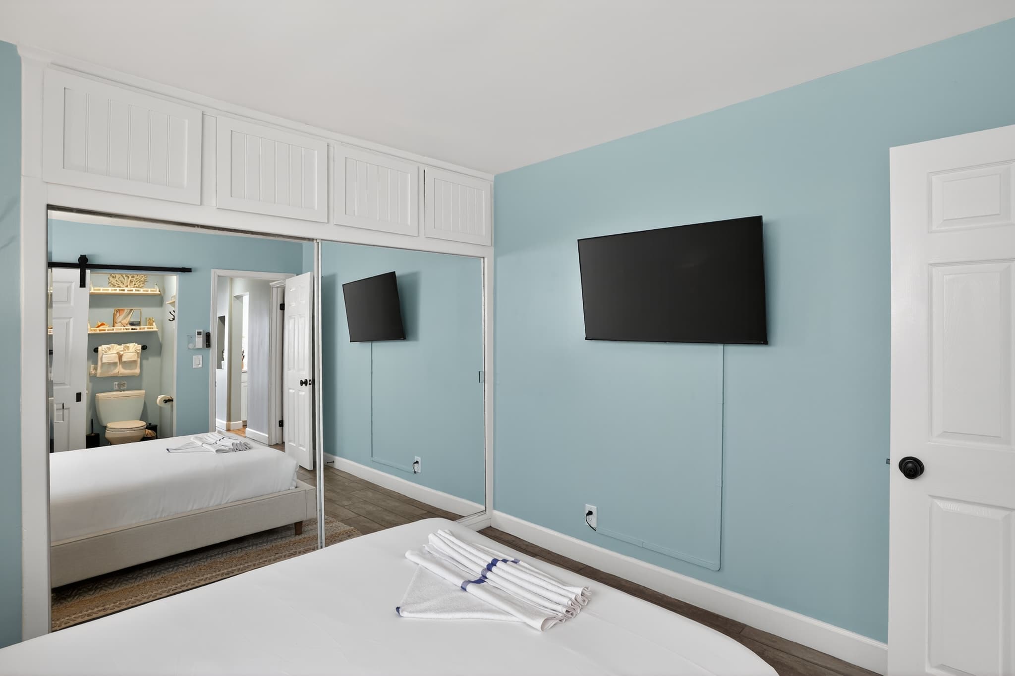 Bedroom with light blue walls and wood-look tile flooring, featuring a large bed with white linens, a wall-mounted TV, and mirrored closet doors with white upper cabinets.