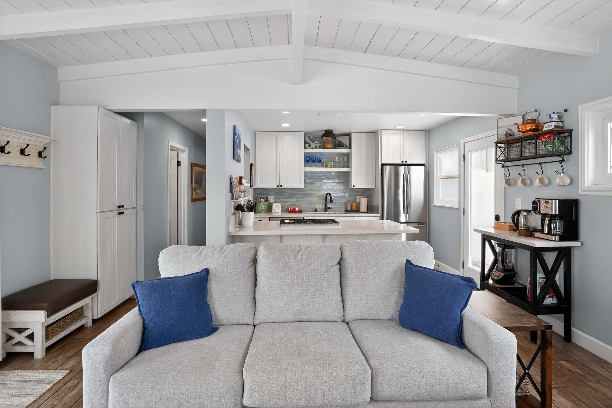 This view from the living room showcases a light gray sofa with blue pillows facing an open kitchen with white cabinetry, light blue backsplash, and a large island with a gas cooktop, all under a white beamed ceiling.