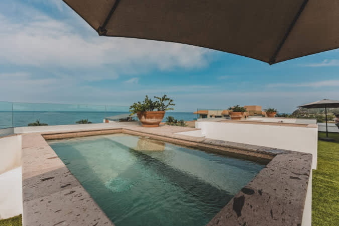 Elevated rectangular swimming pool with stone coping, surrounded by white walls and artificial turf, offering stunning panoramic views of the ocean and distant coastline.