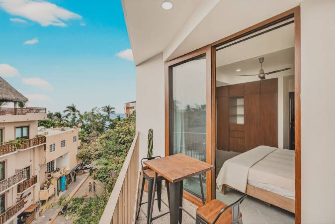 Balcony with a high-top wooden table and two stools, offering a vibrant view of a street below with buildings and a glimpse of the ocean, connected to an adjacent room through a large sliding glass door.