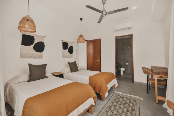 Cozy bedroom with two twin beds, each with a mustard-colored throw, featuring woven wall art and pendant lights, and an en-suite bathroom visible through an open wooden door.