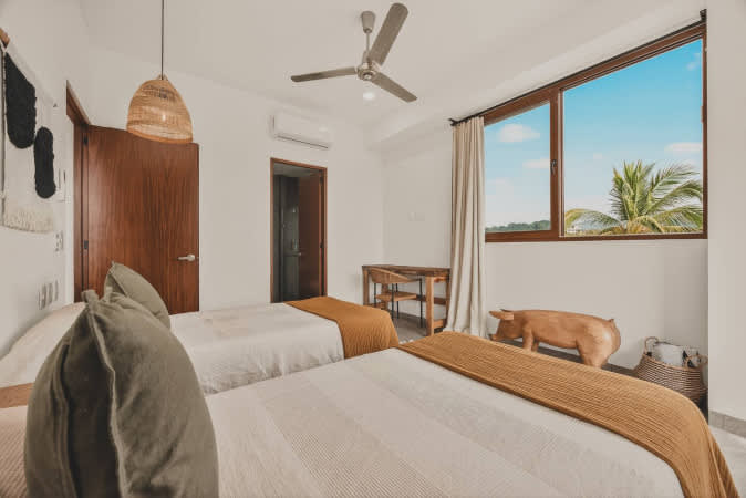 Serene bedroom with two twin beds, a wooden desk and chair, and a large window offering views of palm trees and blue sky, complemented by a decorative wooden pig statue.