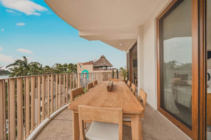 Curved outdoor balcony featuring a large wooden dining table and chairs, offering expansive views of the tropical surroundings and a glimpse of the ocean. Sliding glass doors connect the balcony to the interior living space.