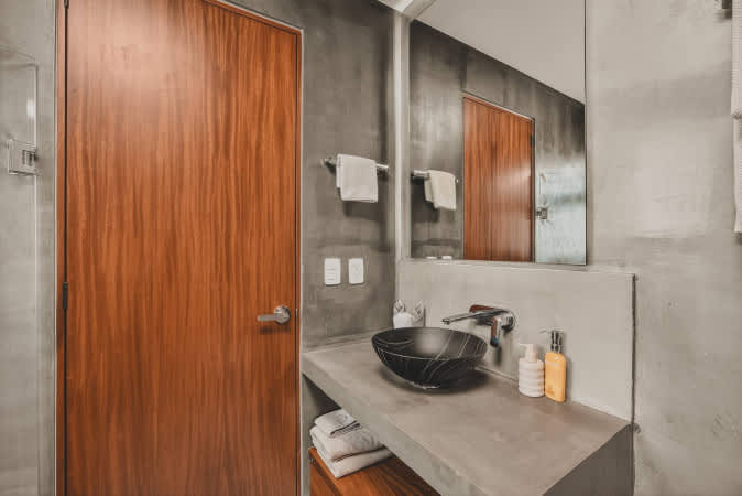 Contemporary bathroom with a black textured vessel sink set on a concrete countertop, featuring a modern faucet and a large mirror, adjacent to a warm wooden door.