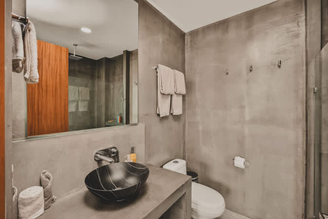 Modern bathroom featuring a dark, textured vessel sink on a concrete vanity, a large mirror reflecting a walk-in shower, and concrete walls with towel racks.