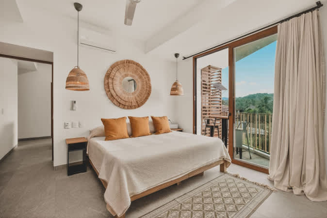 Bright bedroom featuring a large bed with orange accent pillows, two hanging wicker pendant lights, and a circular woven wall decor, with a sliding glass door opening to a balcony with lush green views.