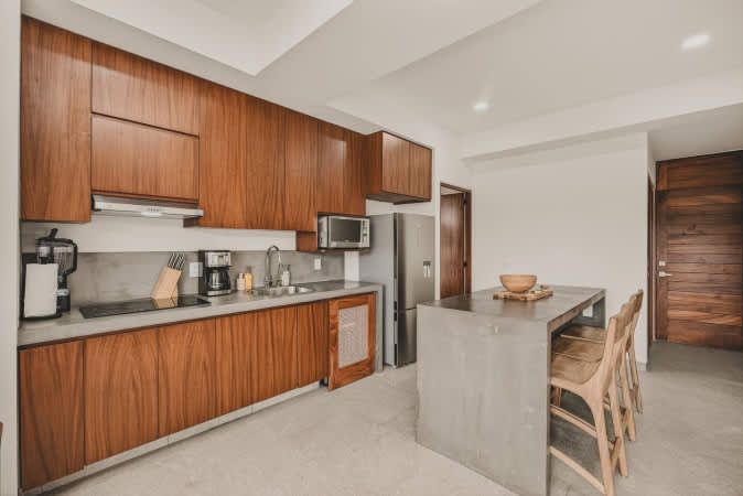 Modern kitchen with dark wood cabinetry, light gray concrete countertops, and stainless steel appliances including a refrigerator, microwave, and stovetop. A blender and coffee maker are on the counter.