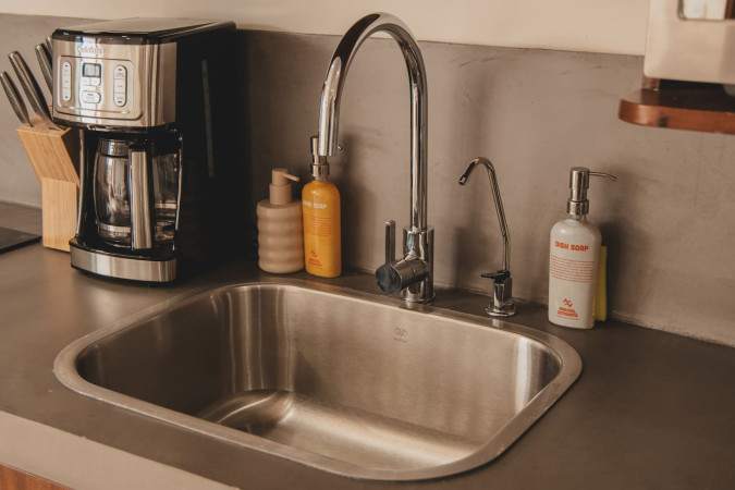 Stainless steel kitchen sink with a gooseneck faucet, accompanied by a coffee maker, knife block, and various soap dispensers on a concrete countertop.