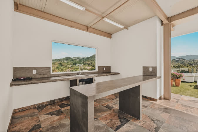 Covered outdoor kitchen area featuring a long concrete bar and matching countertops with a sink, set against white walls and a patterned tile floor, offering a scenic view of green hills through a wide window.
