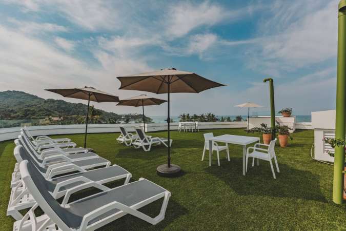 Sunny rooftop deck covered in artificial green turf, furnished with multiple grey and white lounge chairs under large umbrellas, offering expansive views of the ocean and lush green hills.