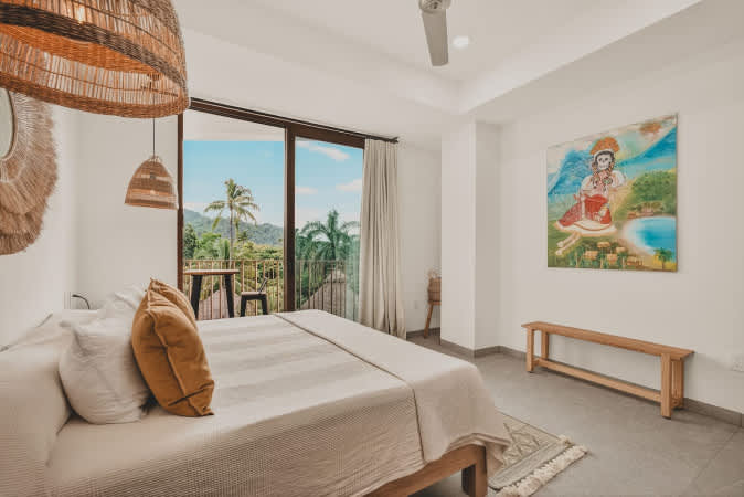 Bright bedroom with a large bed, a woven pendant light, and a large sliding glass door leading to a private balcony with lush tropical views. A colorful painting hangs on the wall.