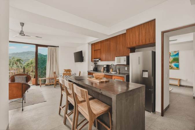 Open-concept living space showcasing a modern kitchen island, a dining table, and a living area visible through large sliding glass doors leading to a balcony with mountain views. A ceiling fan is mounted overhead.