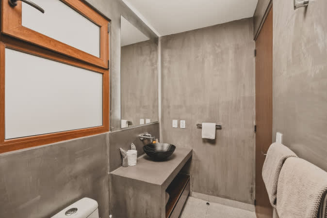 Contemporary bathroom featuring concrete-textured walls, a black vessel sink on a sleek concrete vanity, a large mirror, and a toilet, with a wooden-framed window providing natural light.