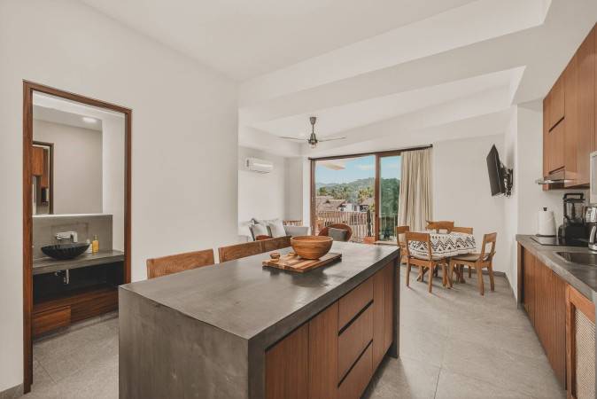 Wide view of the open-concept living area, showing the kitchen island with stools, a dining table with chairs, and a living room with balcony access and mountain views. A half bathroom is visible to the left.