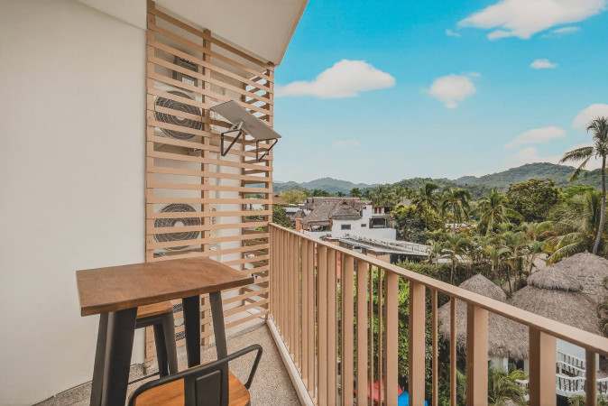 Outdoor balcony space with a small wooden high-top table and two chairs, featuring slatted wooden privacy screens and a scenic view of lush green hills and distant buildings under a clear blue sky.