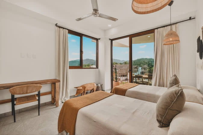 Bright bedroom with two single beds, light bedding, and a woven headboard, featuring large windows and a glass door leading to a balcony with scenic mountain views.