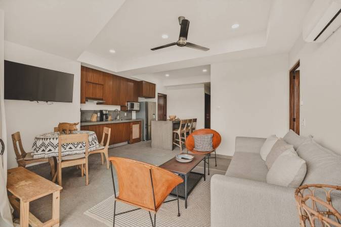 Spacious open-concept living area with a light gray sofa, two orange leather chairs, and a dining table with a patterned cloth. The modern kitchen with dark wood cabinets and a breakfast bar seamlessly integrates into the space.
