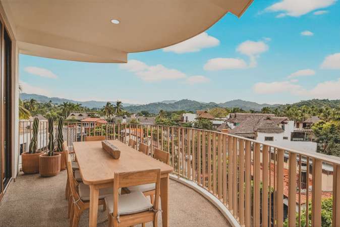 Expansive outdoor balcony featuring a long wooden dining table with seating for eight, perfect for al fresco meals. The balcony offers panoramic views of lush tropical hills, the town below, and distant mountains.