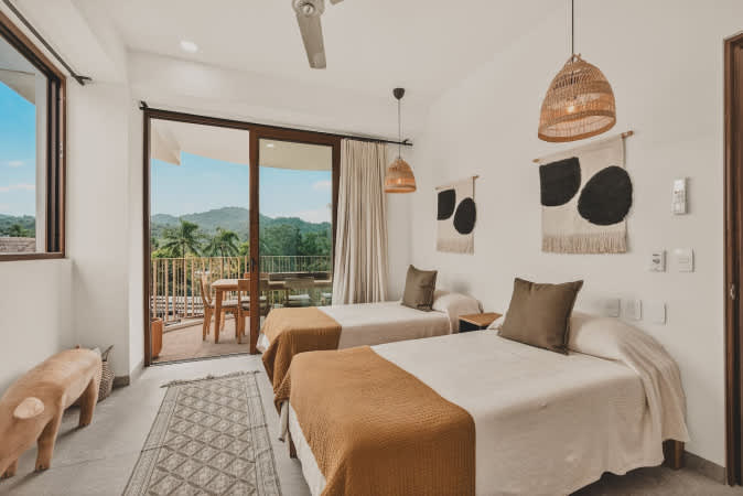 Bright bedroom with two twin beds, each adorned with a mustard-colored throw, featuring woven wall hangings and pendant lights, and a sliding glass door leading to a balcony with lush views.