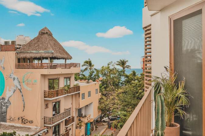Vibrant view from the balcony showcasing the charming architecture of the town, including buildings with thatched roofs and colorful murals, nestled among palm trees. The sparkling blue ocean is visible in the background.