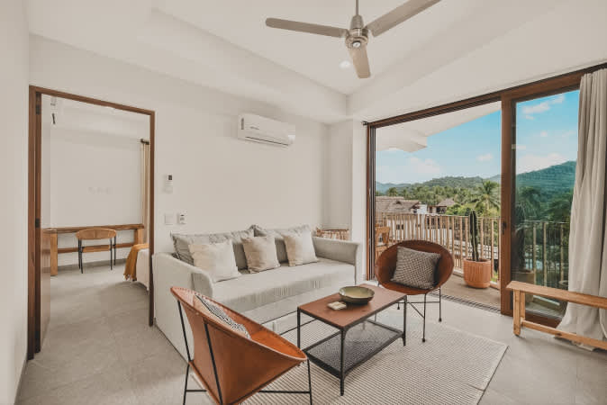 Bright living room featuring a light gray sofa, two brown leather armchairs, and a wooden coffee table, opening to a balcony with lush green mountain views. A ceiling fan and air conditioning unit are visible.