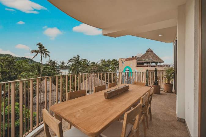 Spacious outdoor balcony with a long wooden dining table and chairs, providing an ideal spot for entertaining or relaxing. The view encompasses a vibrant tropical landscape with palm trees and distant mountains under a clear sky.