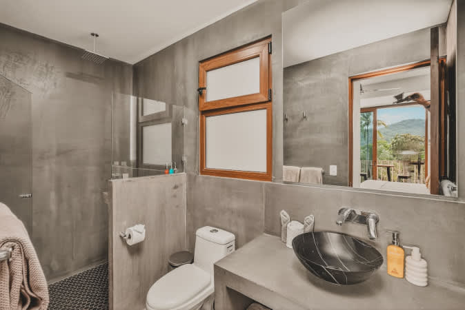 Modern bathroom with concrete-textured walls, a black vessel sink on a minimalist concrete vanity, a toilet, and a walk-in shower with dark hexagonal floor tiles, featuring a wooden-framed window.