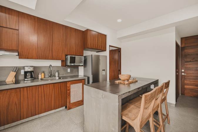 Modern kitchen with dark wood cabinetry and a large concrete island serving as a dining bar with wooden stools. The space opens to a living area and balcony beyond.