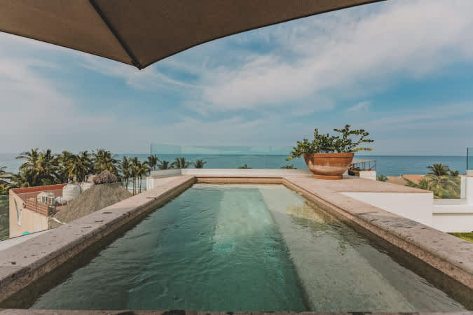 Close-up view of the rectangular swimming pool with clear water and stone edges, positioned on a rooftop to maximize the dramatic ocean and coastal views under a bright blue sky.