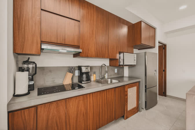 Close-up of the modern kitchen featuring dark wood cabinets, light gray concrete countertops, and integrated appliances including an electric stovetop, microwave, and stainless steel refrigerator. A blender and knife block are on the counter.