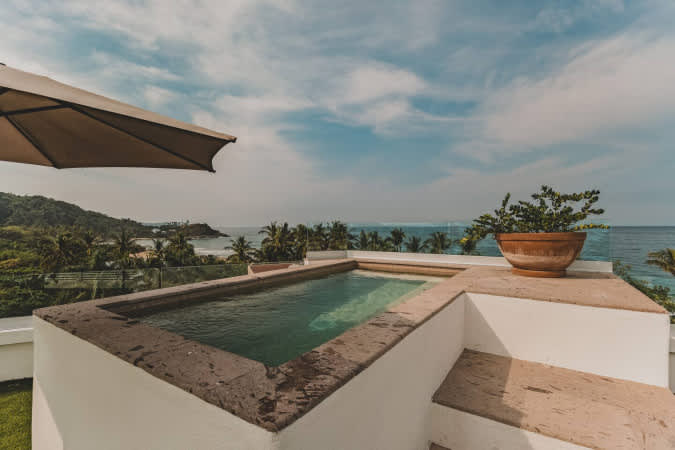 Elevated rectangular swimming pool with stone coping and integrated steps, set on a rooftop with artificial turf, providing expansive views of the ocean and a lush, hilly coastline.