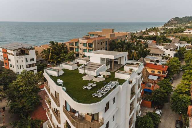 This aerial perspective highlights a contemporary white building featuring a green turf rooftop terrace equipped with lounge seating and small pools. The surrounding coastal town, dense palm trees, and the expansive ocean create a picturesque backdrop.