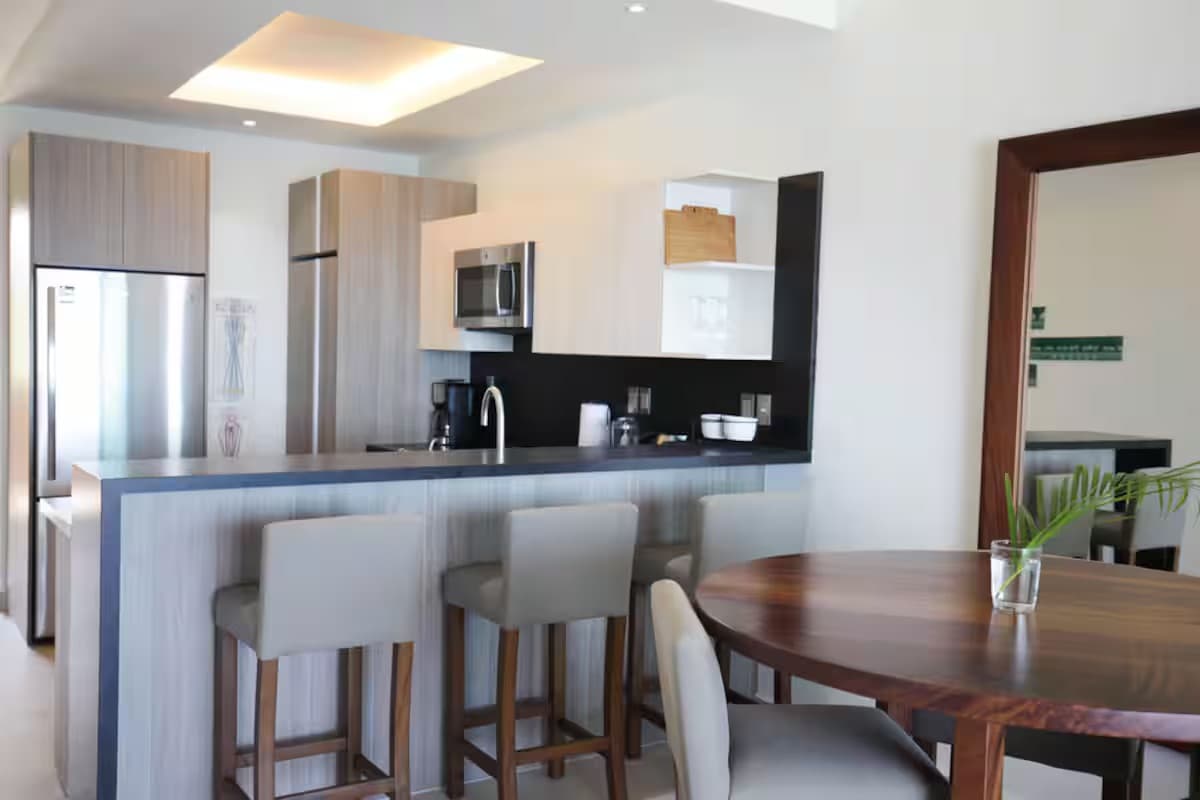 Modern kitchen featuring light wood cabinetry, stainless steel appliances, a dark countertop, and a breakfast bar with three light-colored bar stools, adjacent to a round wooden dining table.